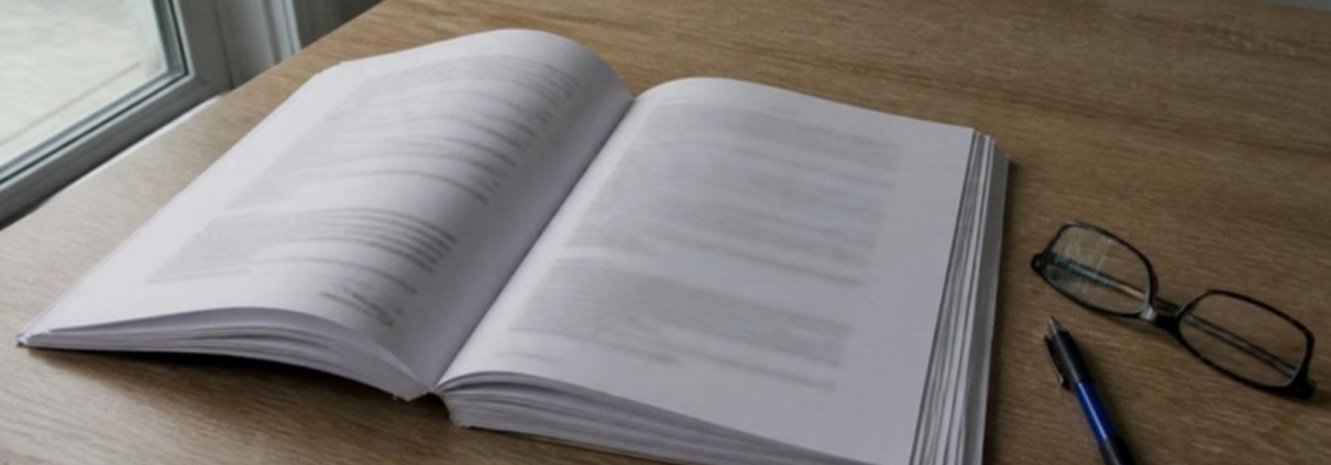 A thick printed document lying open on a wooden table beside a pen and a pair of reading glasses, the text too soft to read.
