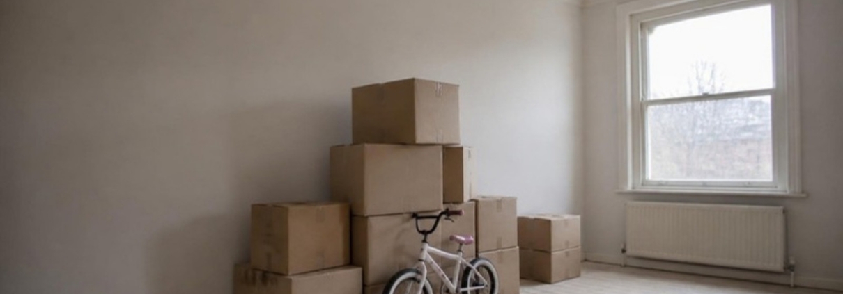 Moving boxes in an empty living room with a child's bicycle beside them.
